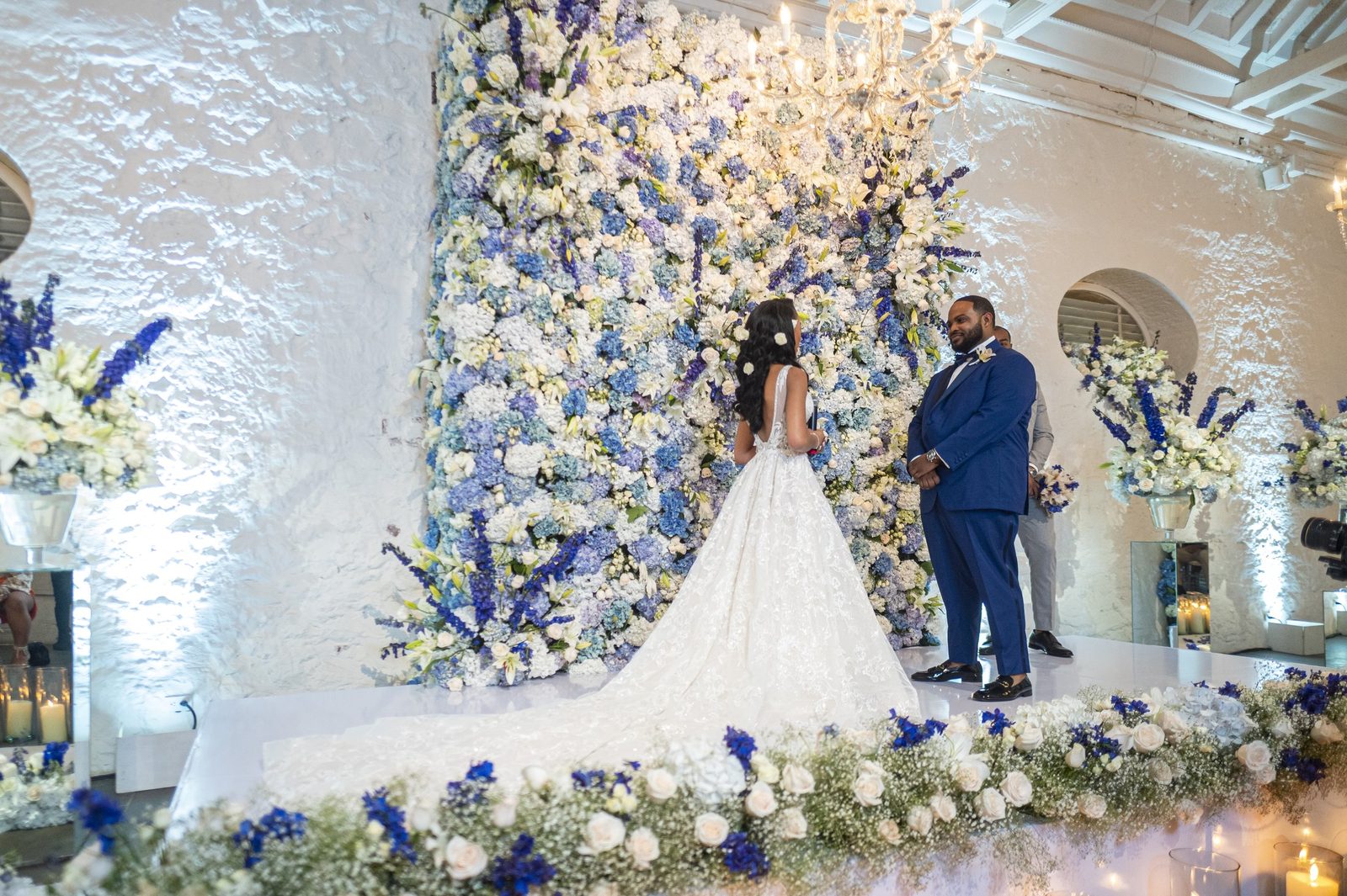 Ceremony in front of a wall of blue and white flowers in a whitewashed colonial hall