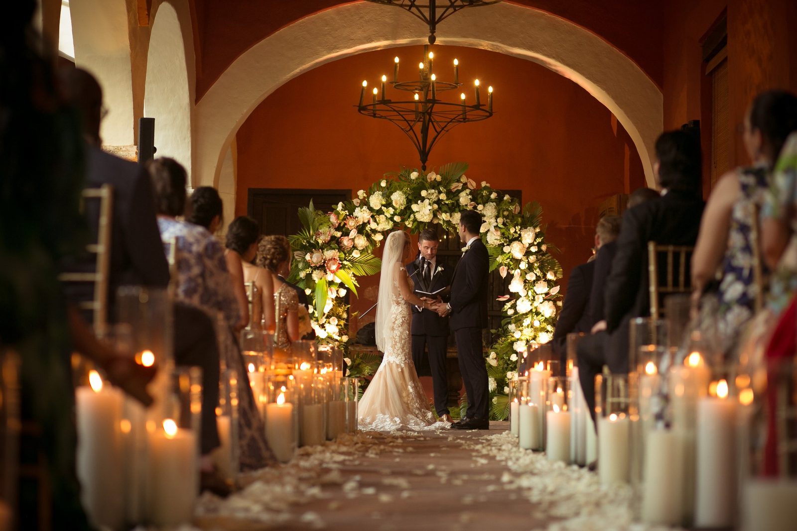 Candlelit aisle leading to a floral arch in a colonial hall with chandeliers