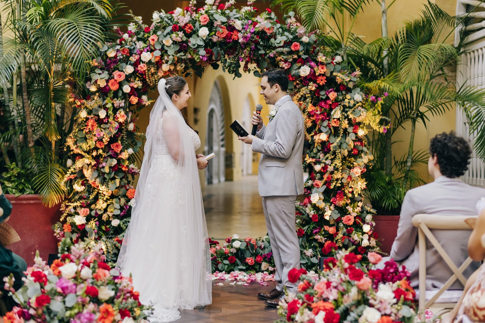 Bride and groom exchanging vows under an arch of roses in a yellow colonial courtyard