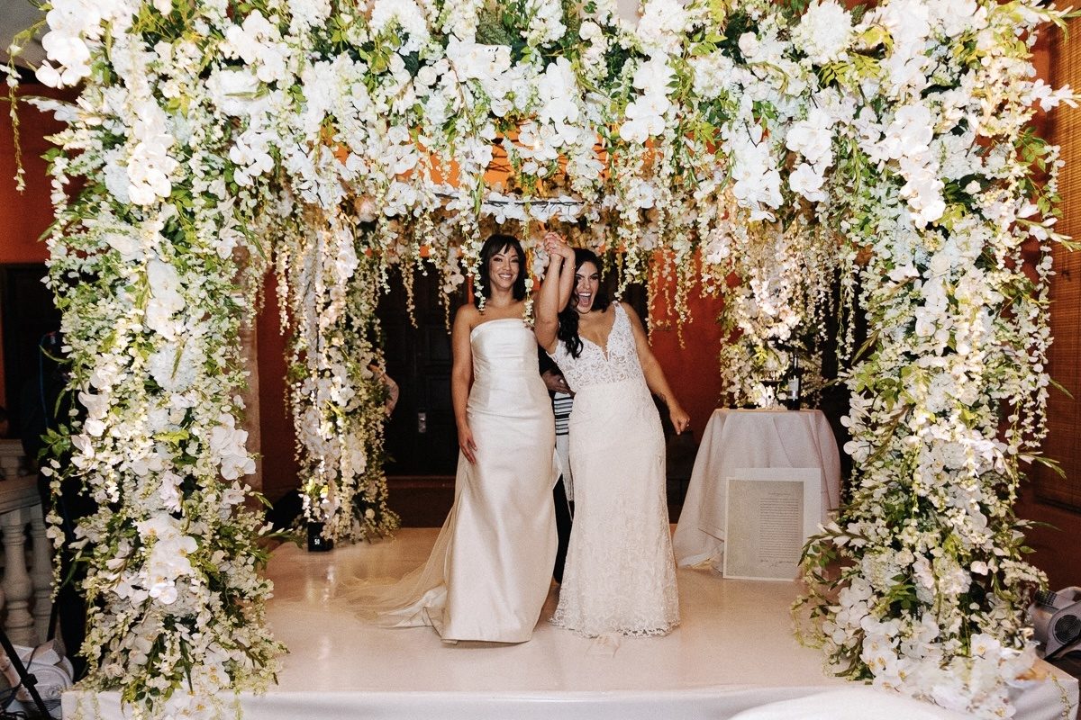 Two brides celebrating under a floral arch of white orchids