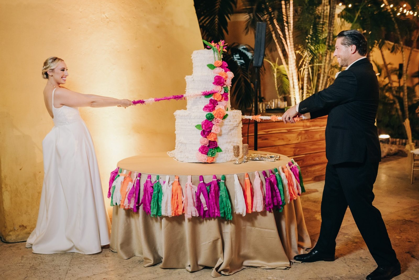 Bride and groom pulling the ribbons of a flower-covered cake piñata